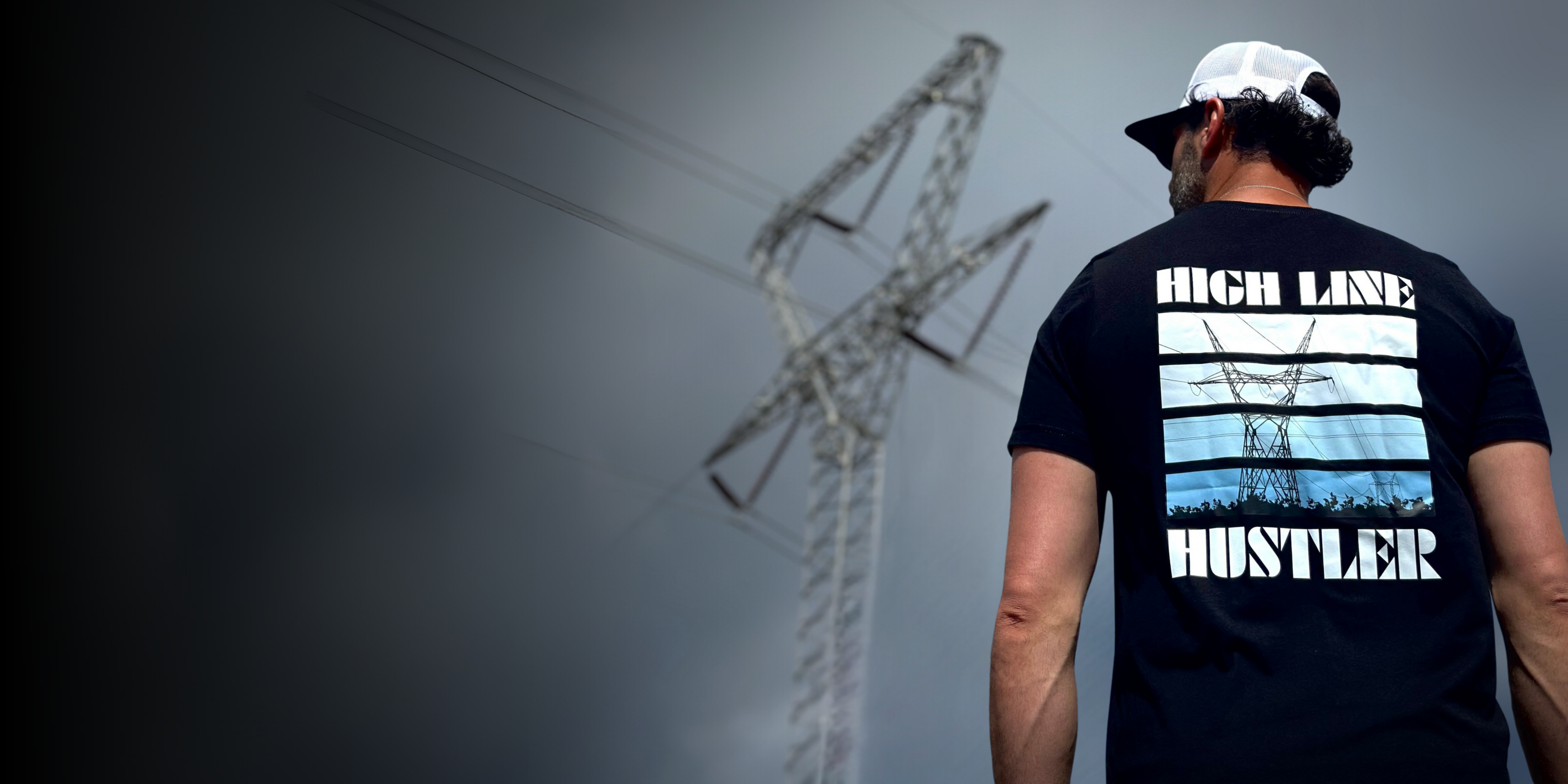 Lineman looking at high voltage tower wearing a black lineman shirt with high line hustler on it
