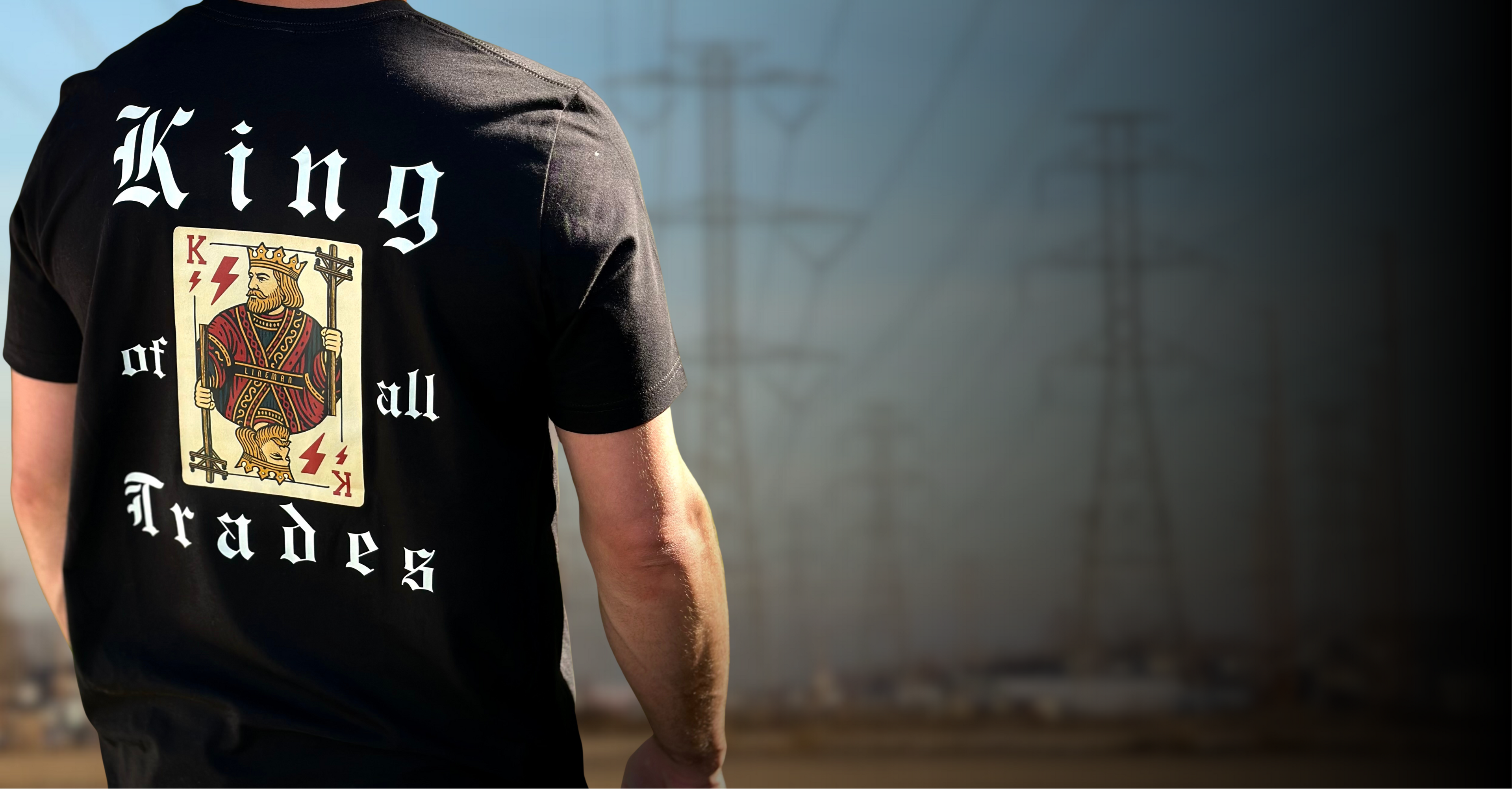 Lineman looking at high voltage towers wearing a black lineman shirt with king of all trades on it.