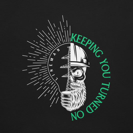 Close up Black lineman shirt with lightbulb, high voltage tower, and skeleton in hard hat. Text that reads lineman and keeping you turned on.